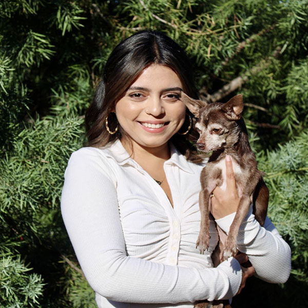 Photo of Nadia Oseguera holding Britney, a brown senior chihuahua in front of a green tree.