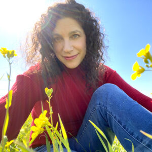 Photo of Sarah in a red sweater and jeans, sitting in a field of yellow flowers