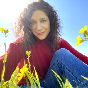 Sarah Reiwitch in a red sweater and blue jeans, sitting amongst yellow flowers