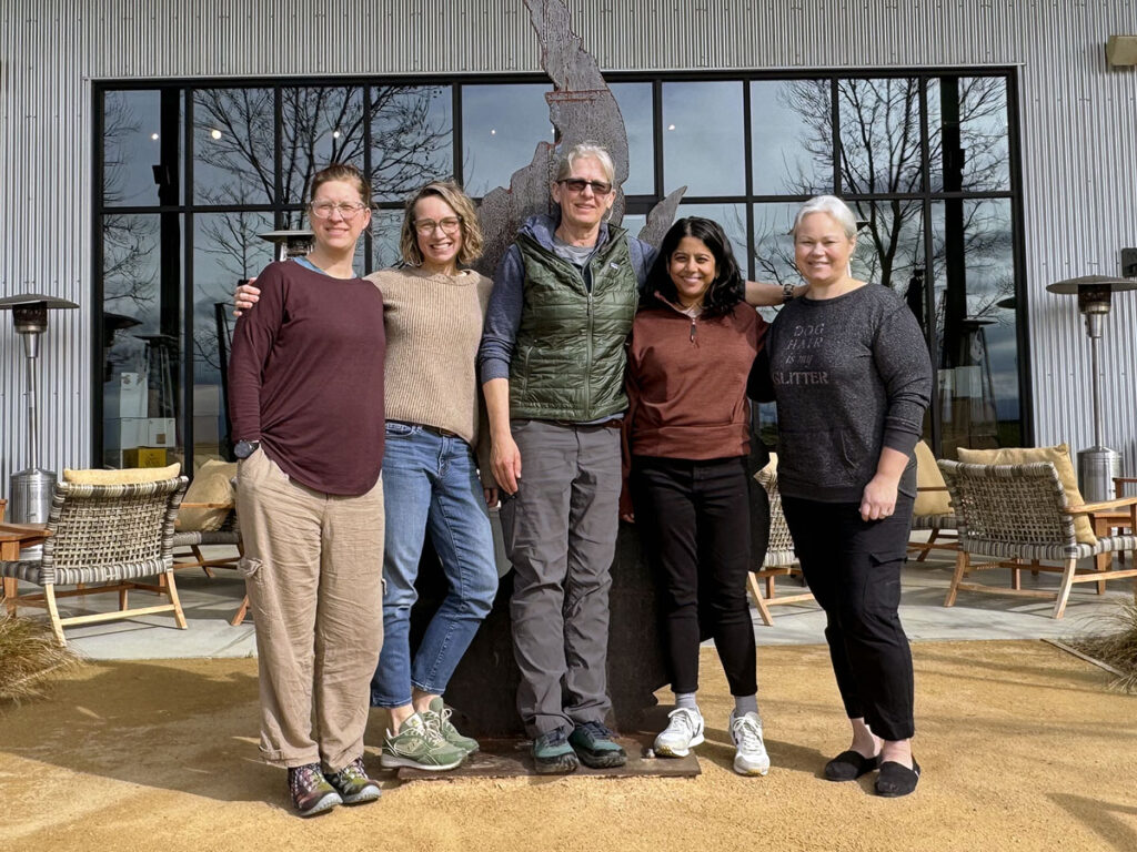Five members of the SMAC team poses on a dirt path in front of a patio with wicker furniture and space heaters.