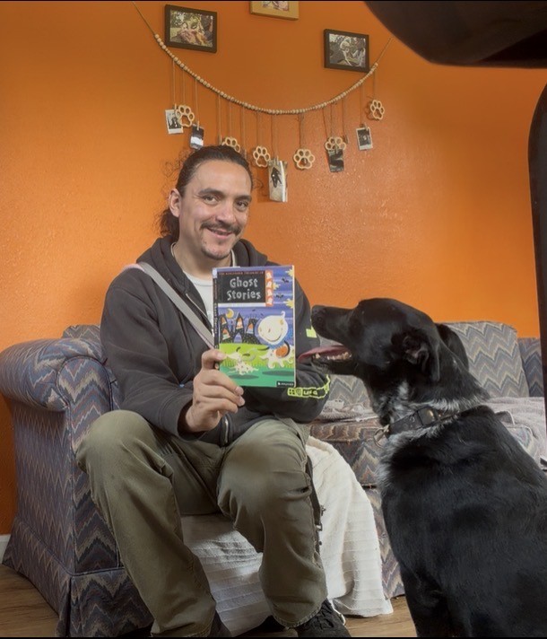 Javier sits on a couch in front of an orange wall holding a book of ghost stories in front of a black dog