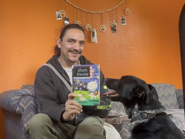 Javier sitting on a couch holding a book of ghost stories in front of a black dog
